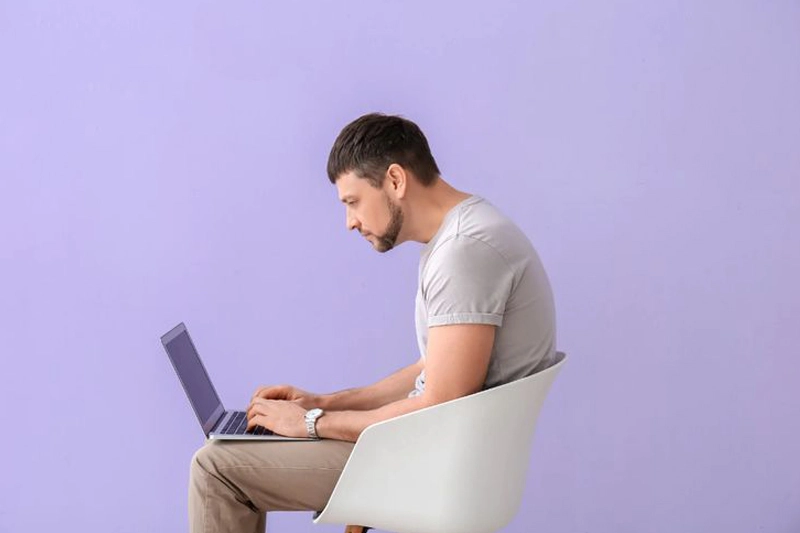 مردی روی صندلی نشسته و لپتاپ را روی پاهایش قرار داده با حالت بد نشستن