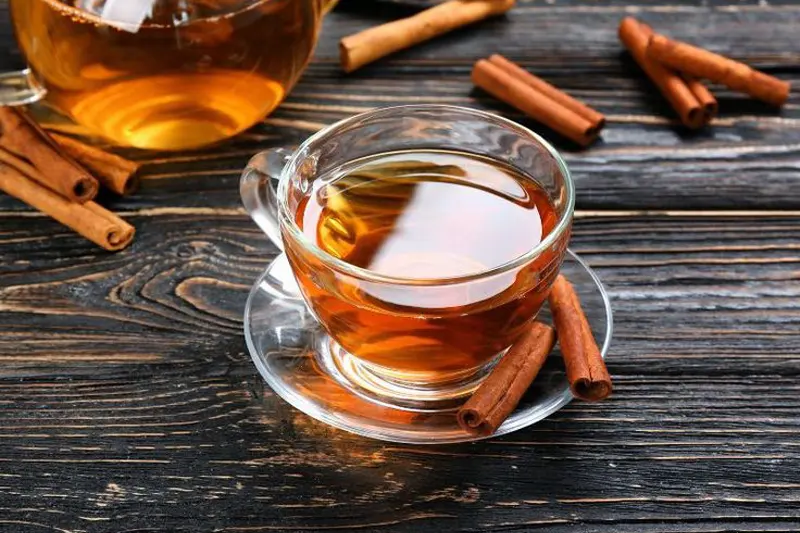 قوری و فنجان پر از دمنوش دارچین با چند رول دارچین اطراف آن