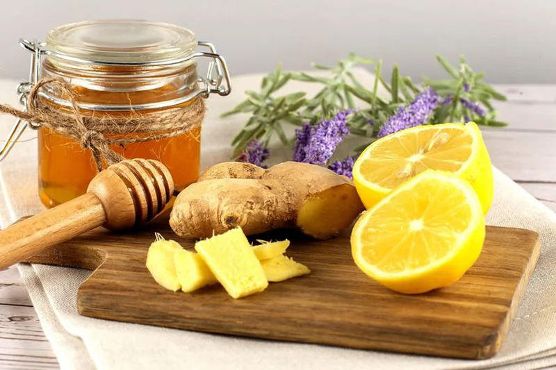 یک شیشه عسل با قاشق چوبی، کنار آن دو نصف لیمو و گیاه زنجبیل، برای تهیه معجون طبیعی ضد سردرد و التهاب. 