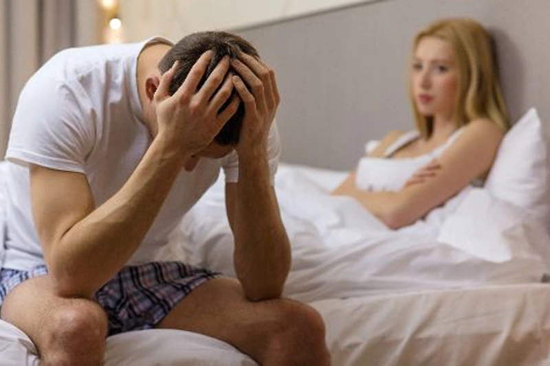 مردی ناامید روی تخت نشسته با شریکش در پسزمینه، قابل استفاده برای کاهش استرس و بهبود کنترل انزال