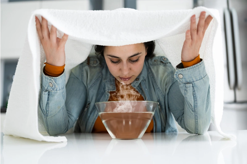 زن زیر حوله در حال انجام بخور نعناع با کاسه بخار برای تسکین سردرد و گرفتگی بینی